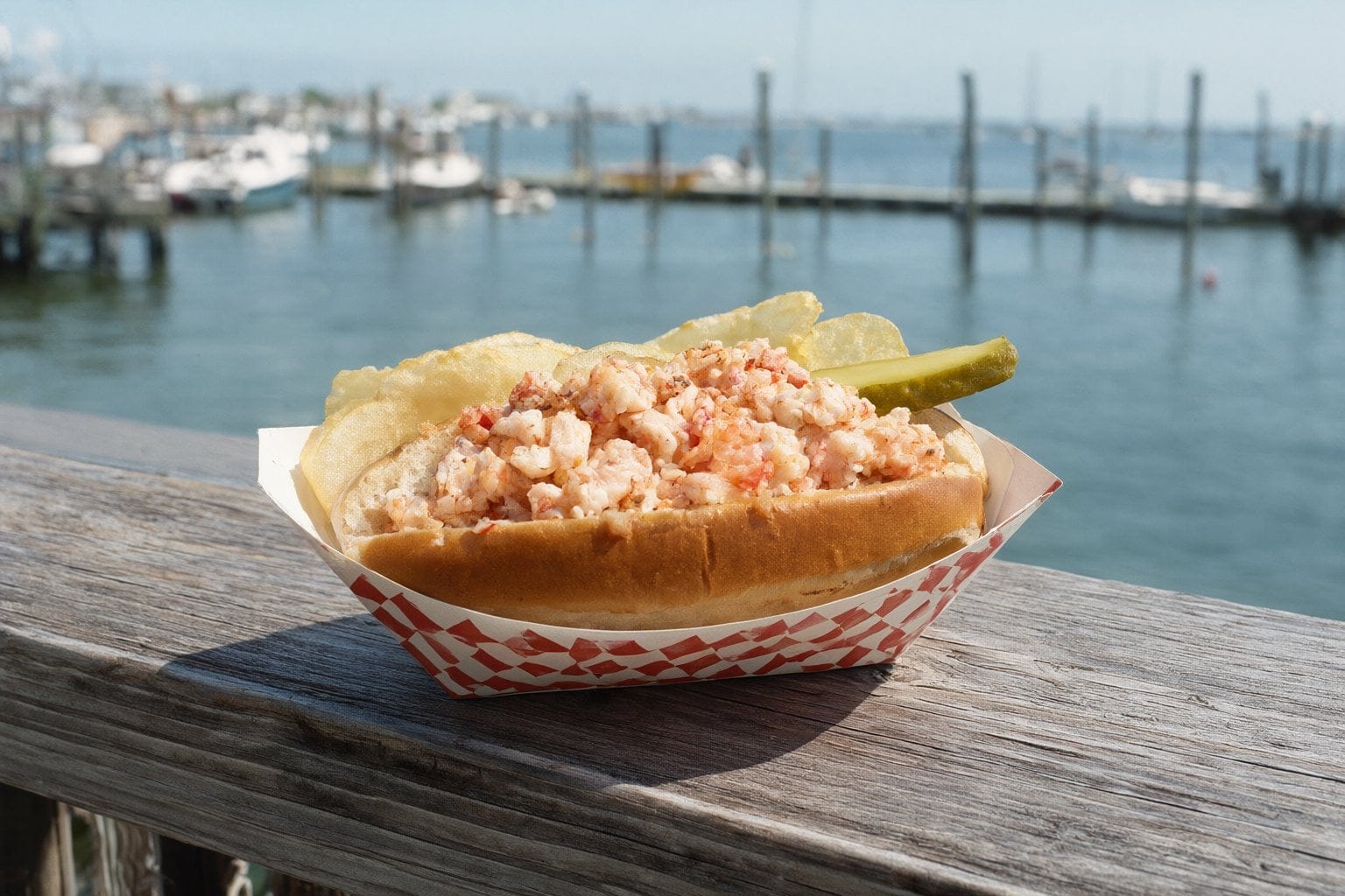 Illustration: a lobster roll with chips and a pickle on a wooden railing above a marina