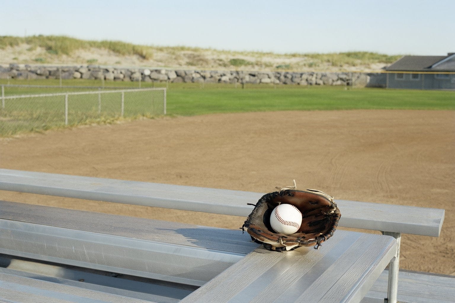 Illustration: a youth baseball glove and ball on aluminum bleachers beside a ball field with dunes behind